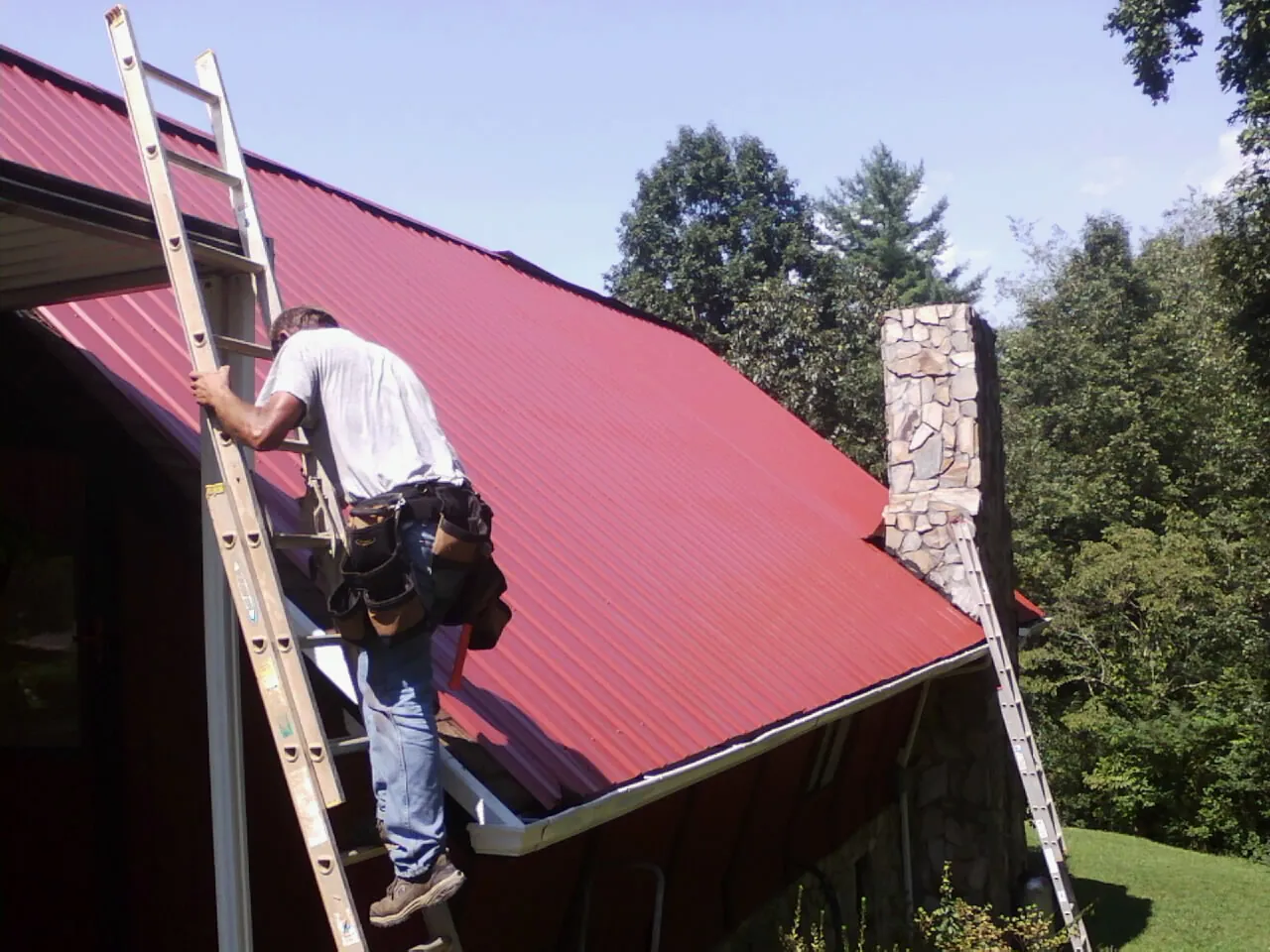 Roofer on a ladder beside a red metal roof and stone chimney — Soffit & Fascia Repair by Roofing Brothers in Woodstock